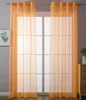 Two orange sheer curtains are hanging in front of a window with a grid pattern. A framed picture of a plant is on the left, and a wooden dresser with a vase and plant is on the right. Produktbild-Vorschau