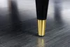 Close-up of the leg of a furniture item, showing a black tapered leg with a gold accent at the bottom, resting on a dark wooden floor. Produktbild-Vorschau