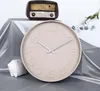 A round, beige wall clock with raised numbers is displayed prominently in the center of the image. Behind it, a vintage-style radio sits on a draped silver fabric. To the left of the clock, a small, textured white decorative object resembling a sheep is vi Anteprima dell’immagine prodotto