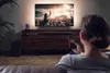 A woman is sitting on a couch, holding a smartphone and looking at a large TV screen displaying a concert. Produktbild-Vorschau