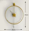 A gold metal wall clock with a pendulum, shown against a white wall. Dimensions are indicated with measurement lines and text. Náhľad obrázku produktu