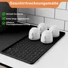 A black drying mat with a textured surface holding four white cups, placed on a countertop with a tiled backsplash in the background. Produktbild-Vorschau