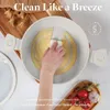 A hand is cleaning a cream-colored frying pan with a sponge, demonstrating its easy-to-clean feature. Ingredients like tomatoes and garlic are arranged in a wooden bowl in the foreground. Produktbild-Vorschau
