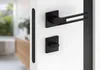 A modern black door handle set mounted on a white door, with a blurred interior background. Produktbild-Vorschau