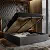 A modern bedroom with a dark gray upholstered bed frame, its storage compartment open, revealing bedding and pillows. The headboard has a tufted design, and the wall behind features a decorative wooden paneling. Náhled obrázku produktu