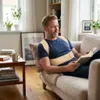 A man is sitting on a couch reading a book while wearing a posture corrector. Anteprima dell’immagine prodotto