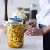 A person's hands are visible on a kitchen counter next to a jar of pickled cauliflower. Other kitchen items like garlic and jars are in the background. Produktbild-Vorschau