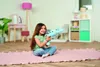 A young girl sits on a pink rug in a room, holding a light blue plush toy. Náhľad obrázku produktu