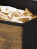 A close-up view of the corner of a fabric storage box, showing the black exterior fabric and a section with a wood-grain pattern. A folded blanket with a tan and white pattern is visible inside the box. Produktbild-Vorschau