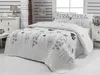 A neatly made bed with a white quilted bedspread featuring a floral pattern in gray, black, and tan. The bed is set in a bedroom with white paneled walls and a bedside table. Podgląd zdjęcia produktu
