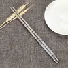 A pair of stainless steel chopsticks rests on a textured gray fabric background, with a few stalks of wheat and a white bowl visible. Produktbild-Vorschau