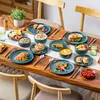 A beautifully set dining table with a full set of blue stoneware dishes, featuring various food items like croissants, fruit, salad, soup, and sandwiches, arranged on a wooden table with chairs in the background. Náhled obrázku produktu
