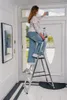 A woman is cleaning a window above a door while standing on a stepladder. Produktbild-Vorschau
