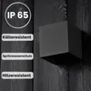 A black cube-shaped outdoor wall light mounted on a dark wooden wall, with text overlays describing its features. Voir les images du produit