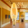 A vintage-style LED bulb is placed in the foreground of a room that appears to be a museum or gallery, with artwork on the walls and chandeliers overhead. Produktbild-Vorschau