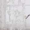 A close-up view of a sheer curtain with a baroque-style pattern, partially covering a window. A white vase with a green plant and a yellow flower is visible to the right. Produktbild-Vorschau