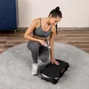 A woman in workout clothes is kneeling on a rug next to a vibration plate, holding a remote control. Produktbild-Vorschau
