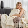 A woman is sitting in a chair, wrapped in a soft blanket, looking away from the camera with a smile. Produktbild-Vorschau