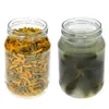 Two glass jars are shown side-by-side against a white background. The left jar is filled with dry pasta, a mix of green and yellow macaroni. The right jar contains pickles submerged in brine. Podgląd zdjęcia produktu
