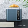 A blue toaster with a croissant on top, placed on a black surface with a gray, textured background. A white cloth is visible in the foreground. Voir les images du produit