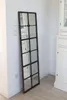 A tall, rectangular mirror with a dark frame, resembling a window, leans against a white wall next to a white cabinet. Produktbild-Vorschau
