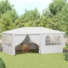 A white party tent set up in a grassy backyard with trees in the background. A table and chairs are visible inside the tent. Voir les images du produit