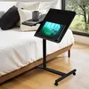 A mobile desk with a laptop stand is placed next to a bed, with a window in the background showing trees. Náhľad obrázku produktu