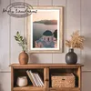 A framed picture of a church on a cliff overlooking the sea is displayed on a wooden shelf unit. Two vases, one with greenery and the other with dried grass, are placed on top of the shelf. A woven basket and a decorative pot are inside the shelf. Produktbild-Vorschau