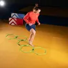 A woman is performing an agility exercise on a yellow mat in a gym, stepping through a series of green hexagonal agility rings. Two stacked boxes with red and blue markings are visible in the background. Náhled obrázku produktu