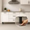 A close-up of a black adjustable furniture leg being installed under white kitchen cabinets. Náhled obrázku produktu