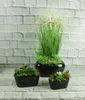 Three black ceramic planters with artificial plants are arranged on a surface in front of a white brick wall. Náhľad obrázku produktu