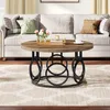 A round coffee table with a wood-grain top and a black metal base with circular accents, placed in a living room setting in front of a sofa. Podgląd zdjęcia produktu