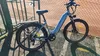 An electric bicycle is parked next to a chain-link fence on a sunny day. Produktbild-Vorschau