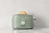 A green Haden toaster with a slice of toast in it, placed on a marble countertop against a white and gray textured background. Náhled obrázku produktu