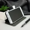 A smartphone in a case, propped up on a surface, with a plant and a pen in the background. Produktbild-Vorschau
