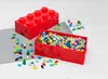 Two red LEGO storage boxes, one closed and shaped like a large LEGO brick, and another open and filled with colorful LEGO bricks, are displayed on a light gray mat and a white surface. Voir les images du produit