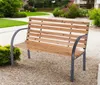 A wooden garden bench is placed on a gravel path in a garden setting with green grass and bushes. Produktbild-Vorschau