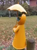 A yellow garden figurine of a frog wearing a raincoat and holding an umbrella, standing in a garden with fallen leaves. Produktbild-Vorschau