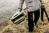 A person walks along a rocky shore next to a body of water, carrying a portable pizza oven and a tripod. Produktbild-Vorschau