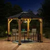 A wooden gazebo with a black roof is set up on a paved patio at night, illuminated by interior lighting. It contains a small table and two chairs, with plants and decorative items around it. Produktbild-Vorschau