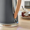 Close-up of a gray electric kettle with a wooden handle, showing a blue indicator light at the base. A jar of jam and some toast are visible in the background. Produktbild-Vorschau
