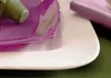 A close-up view of a white plate with a purple plate on top, set on a green textured surface. A purple napkin is visible in the background. Anteprima dell’immagine prodotto