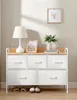 A white fabric dresser with a wooden top and five drawers, placed against a light gray wall. A lamp, a framed picture, and a vase with greenery are on top of the dresser. Shoes and slippers are on the floor below. Produktbild-Vorschau