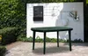 A dark green plastic garden table is set on a patio in front of a white wall with a chalkboard and garden tools. Voir les images du produit