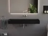 A modern bathroom sink with a black matte finish, mounted on a gray wall, with a large illuminated mirror above it reflecting a plant and chairs. Produktbild-Vorschau