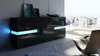 A modern black sideboard with blue LED lighting is placed in a living room with a large window overlooking a city skyline. Produktbild-Vorschau