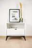 A white nightstand with a drawer and an open shelf, decorated with a framed picture and a vase with dried flowers. Produktbild-Vorschau