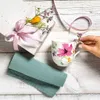 A Villeroy & Boch Mariefleur Basic mug with a floral design, placed on a wooden surface next to a gift-wrapped box and a folded napkin. A hand is shown gently touching a sprig of flowers near the mug. Podgląd zdjęcia produktu