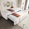 A white sofa bed with a pull-out trundle bed, set up in a room with framed pictures on the wall and a washing machine in the background. Produktbild-Vorschau