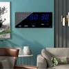 A digital clock with calendar and temperature display mounted on a teal wall, with furniture and decor in the foreground. Produktbild-Vorschau
